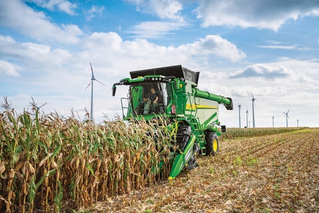 Mähdrescher mit Maiserntevorsatz bei der Maisernte auf einem Feld mit Windkraftanlagen im Hintergrund