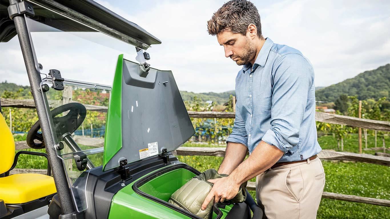 Eine Person legt eine gepolsterte Tasche in das vordere Staufach eines grünen John Deere Gator GS, der draußen in der Nähe eines Holzzauns und eines Weinbergs geparkt ist.