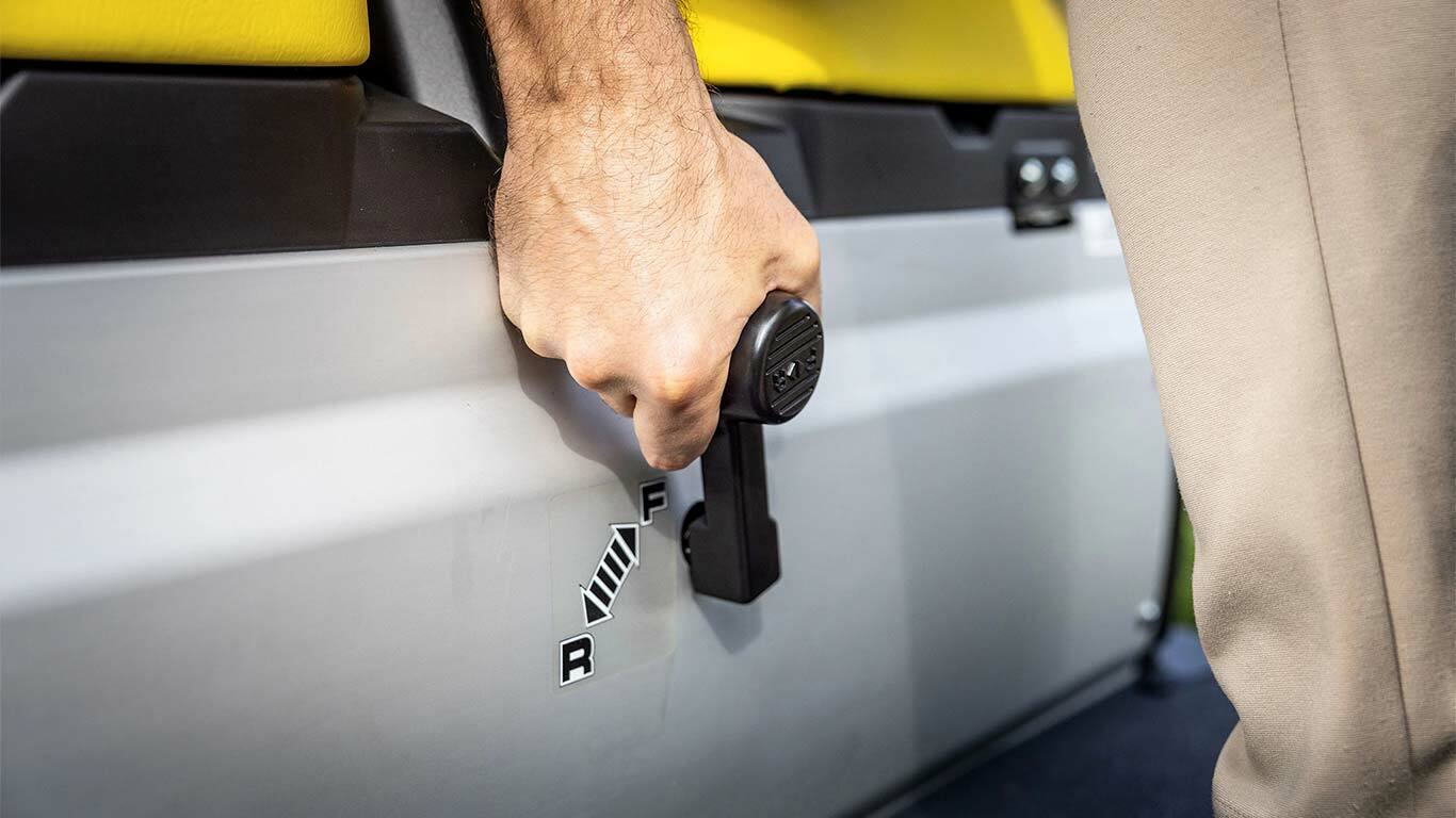 Nahaufnahme einer Hand, die den Schalthebel eines John Deere Gator GS bedient, mit den Vorwärts- und Rückwärtspositionen neben den gelben Sitzen.