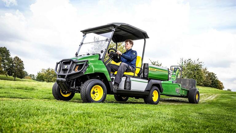 Grünes John Deere Gator Electric Nutzfahrzeug zieht Geräte über ein grasbewachsenes Feld unter teilweise bewölktem Himmel.
