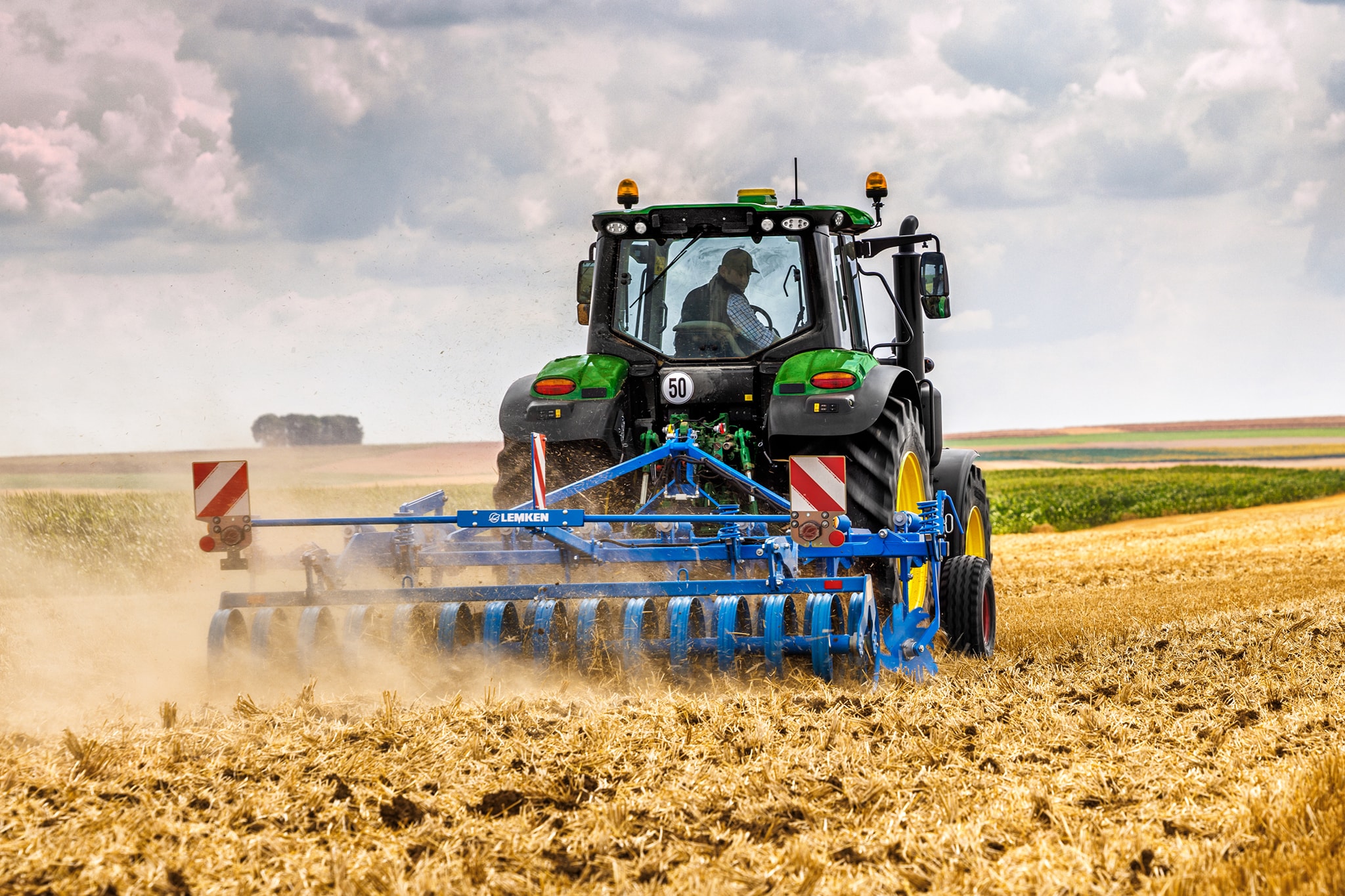 John Deere Traktoren der Serie 6M bearbeitet ein abgeerntetes Feld mit einer Scheibenegge; Rückansicht unter staubigen Feldbedingungen.