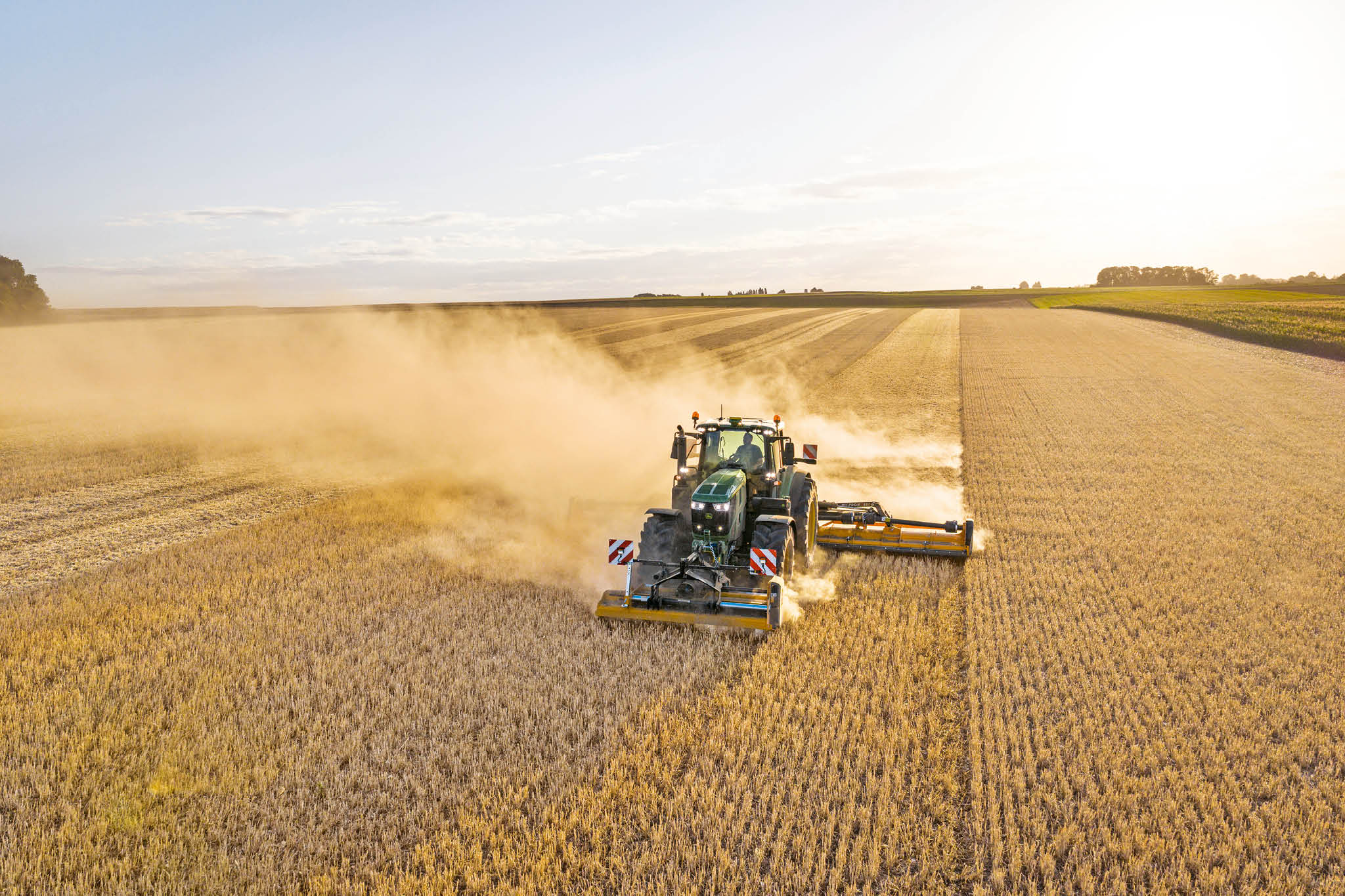 Un tracteur John Deere de la série 6M tractant un épandeur à lisier sur une route de campagne bordée d’herbe et de champs.