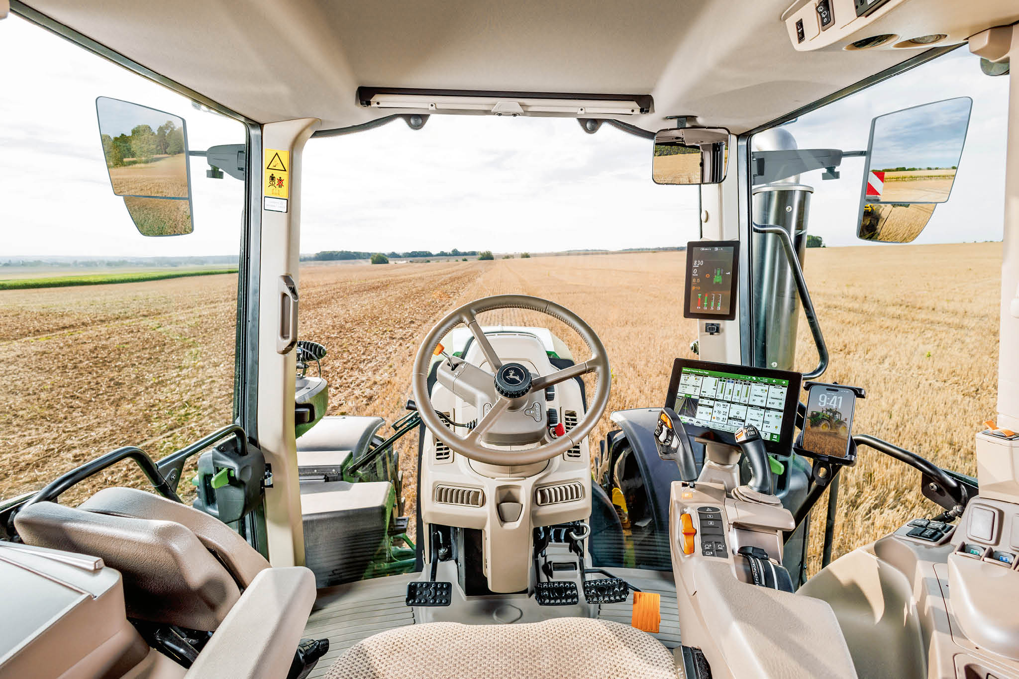 Vue intérieure de la cabine d’un tracteur John Deere de la série 6M, montrant le volant, les consoles de commande et un champ de chaume devant.