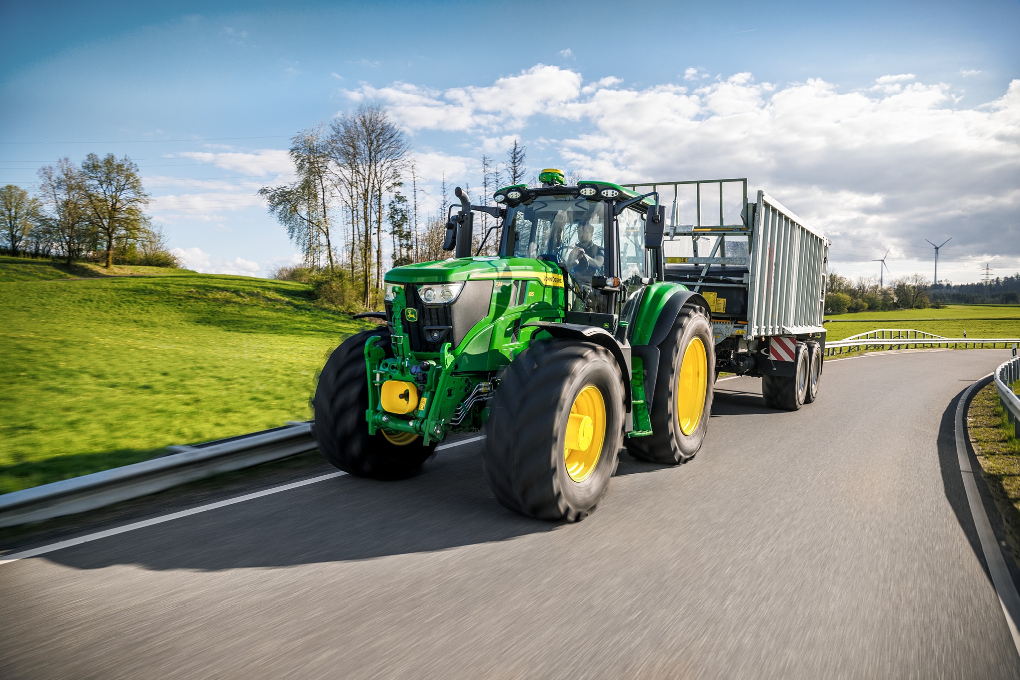 Un tracteur John&nbsp;Deere de la série&nbsp;6M tractant une remorque à ridelles hautes sur une route de campagne goudronnée.