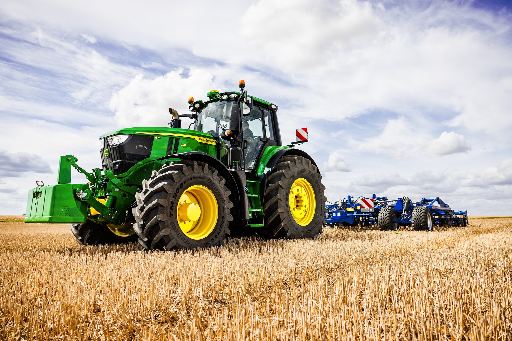 Un tracteur John&nbsp;Deere de la série&nbsp;6M tire un équipement de travail du sol lourd à travers un champ de chaume sous un ciel nuageux.