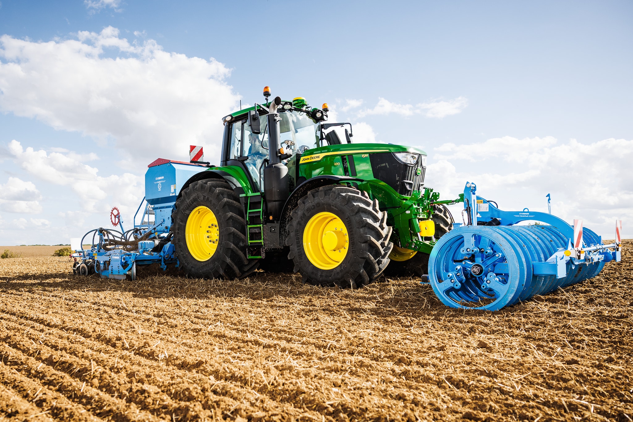 Un tracteur John&nbsp;Deere de la série&nbsp;6M en train de labourer un champ, doté d’un grand équipement à rouleau bleu.