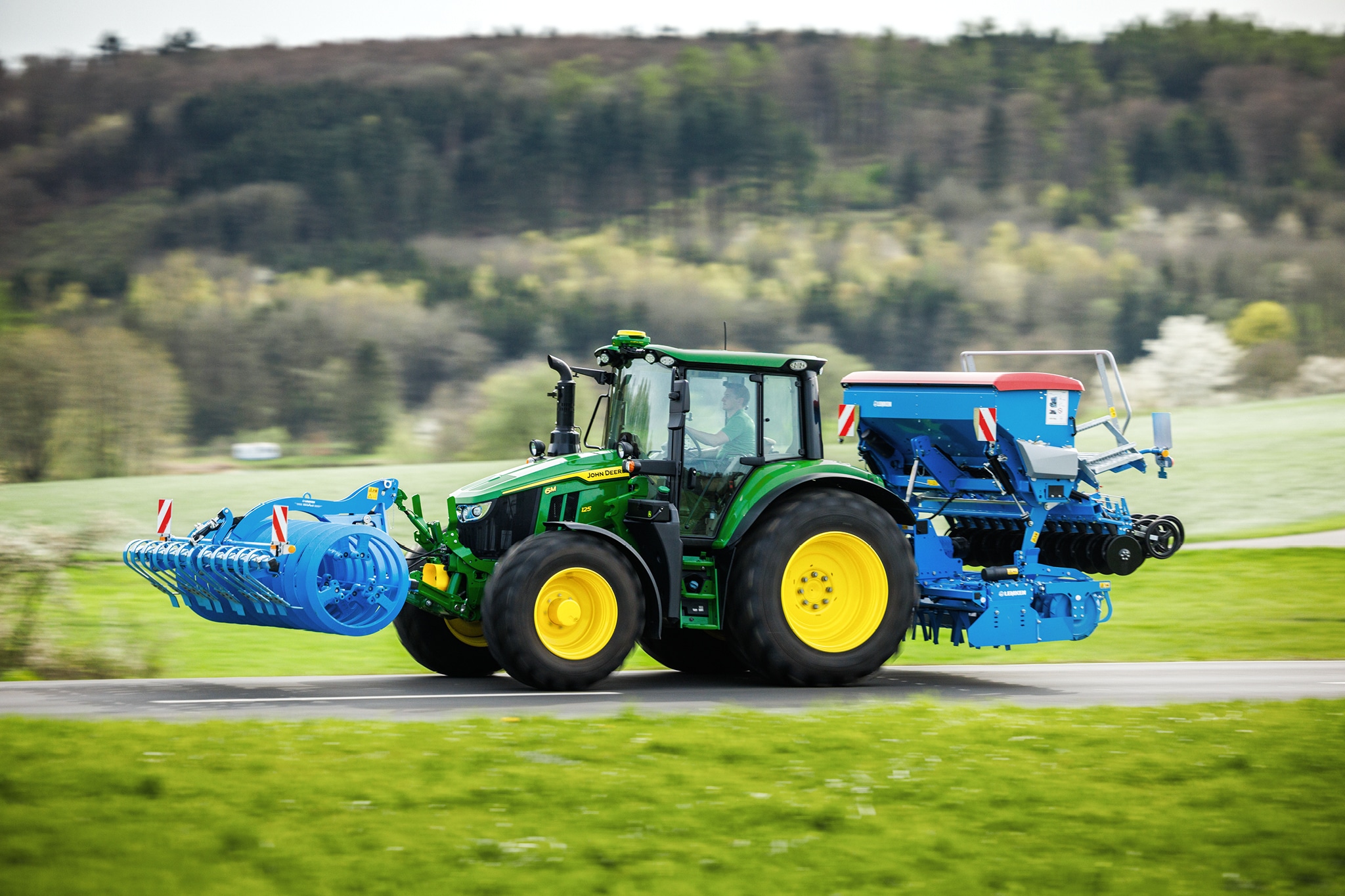John Deere Traktoren der Serie 6M transportieren ein Frontanbaugerät und eine Hecksämaschine entlang einer Landstraße.
