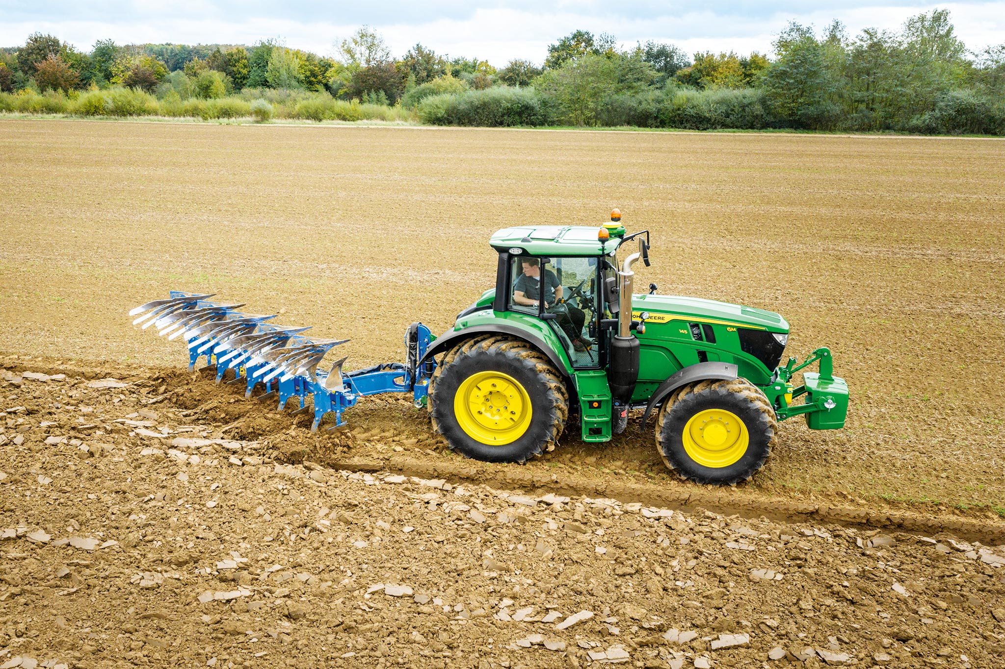 Un tracteur John&nbsp;Deere de la série&nbsp;6M dans un champ labouré avec un équipement de travail du sol monté à l’arrière, sous un ciel nuageux.