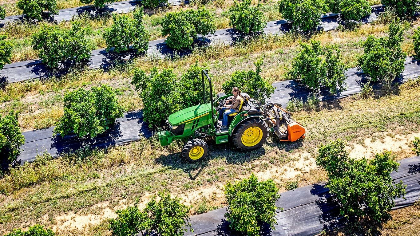 Grün-gelber John Deere Traktor 5075EN bei der Arbeit im Obstgarten, der den Boden zwischen mit Bodenmatten abgedeckten Obstbaumreihen bearbeitet.