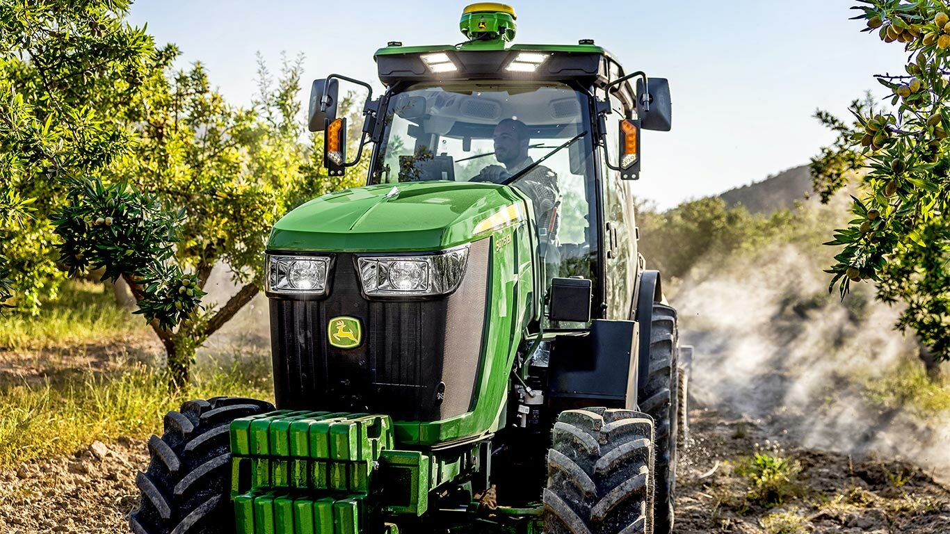 Grün-gelber John Deere Traktor 5075EN, der auf einem Feldweg durch einen Obstgarten fährt, umgeben von Obstbäumen und Sonnenlicht.