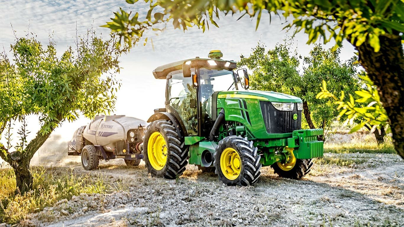 Grün-gelber John Deere Traktor 5090EN, der eine Feldspritze an einem sonnigen Tag durch einen Obstgarten zieht.