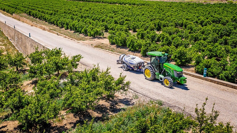 Grün-gelber John Deere Traktor 5090EN, der eine Feldspritze auf einer asphaltierten Straße zwischen großen Obstbaumreihen unter klarem blauem Himmel zieht.