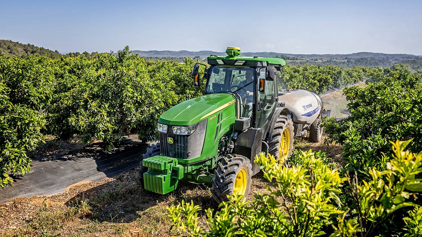 Grün-gelber John Deere Traktor 5105EN, der eine Feldspritze in einem üppigen Obstgarten zieht, mit sanften Hügeln im Hintergrund.