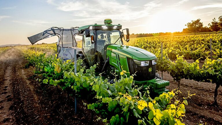 Grün-gelber John Deere Traktor 5105EN, der bei Sonnenuntergang in einem Weinberg arbeitet und die Pflanzen zwischen den Rebenreihen besprüht.