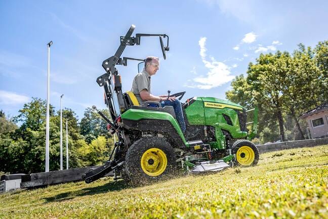 Fahrer, der mit einem John Deere Kompakttraktor und einem Zwischenachsmähwerk einen Grashang mäht