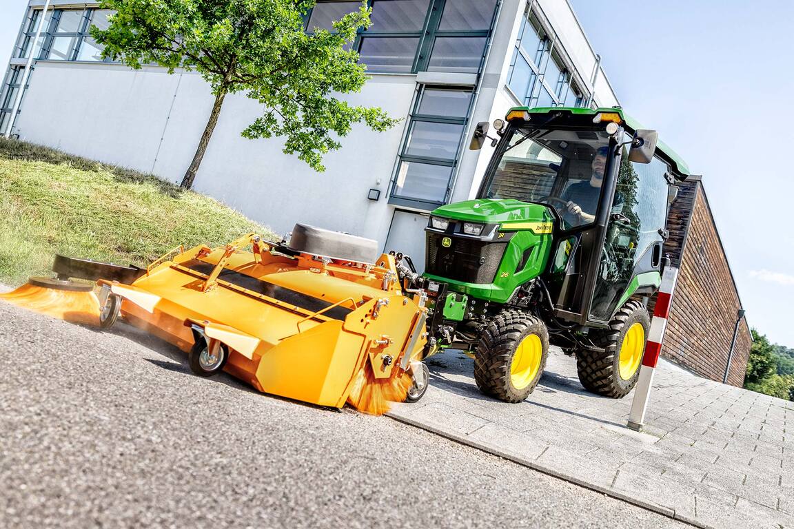 John Deere Kompakttraktor mit geschlossener Kabine und Frontkehrmaschine, der neben einem modernen Gebäude Laub unter Bäumen kehrt