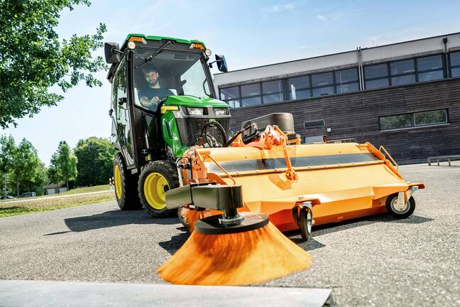 John Deere Kompakttraktor mit geschlossener Kabine und Frontkehrmaschine, der auf einem gepflasterten Bereich Laub unter Bäumen kehrt