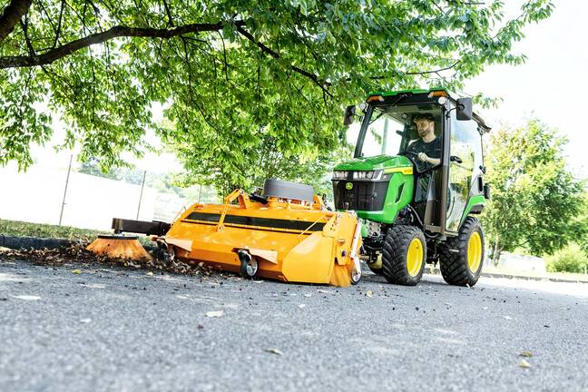 John Deere Kompakttraktor mit geschlossener Kabine und Frontkehrmaschine, der Laub unter Bäumen kehrt
